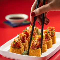 A close-up action shot of chopsticks picking up a piece of crispy tuna sushi for Harajuku Tokyo Cafe. The bright red background makes the image pop.