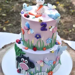 A close-up of an incredibly detailed two-tier birthday cake for a woodland-themed party. The cake features edible butterflies, bees, ladybugs, and a bunny topper, perfectly matching the event's decor.