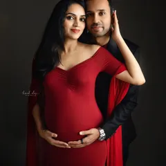 An elegant couple's maternity portrait against a dark grey backdrop. The woman's vibrant red gown and the intimate pose create a dramatic and romantic fine-art photograph.