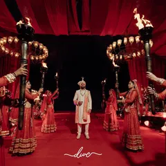 A grand and royal groom entry. He stands confidently in the center of a red-carpeted stage, flanked by performers holding torches, ready for his wedding.