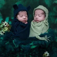 These newborn twins are styled in adorable knit bonnets, one in navy blue and one in sage green, nestled together in a dark, moody, forest-themed setup.