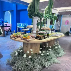 A beautifully composed grazing table set against a striking blue backdrop. The arrangement features tall floral centerpieces, tiered platters, and a variety of gourmet foods, all resting on a custom table surrounded by a carpet of flowers.