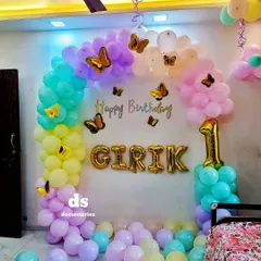 A whimsical butterfly theme for a first birthday, featuring a pastel rainbow balloon arch adorned with golden butterfly stickers. The child's name and age are highlighted with large foil balloons.
