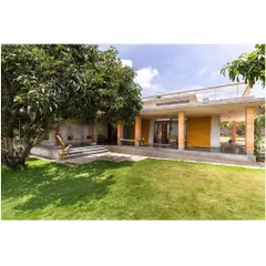 The exterior of "Aamra," where the L-shaped structure opens up to a lush lawn and mango orchard. The design is based on the 'Jagli katte' concept, a traditional verandah space that runs along the boundary, creating numerous semi-open areas for leisure.