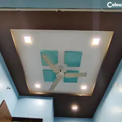 This bedroom ceiling features a square-in-square design with a contrasting color palette. The central section incorporates textured panels and is framed by recessed lighting, creating a neat and centered look around the fan.