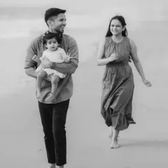 A black and white photograph of a family walking along the shore. The father carries their child while the mother walks alongside, creating a timeless image of a family on the move.
