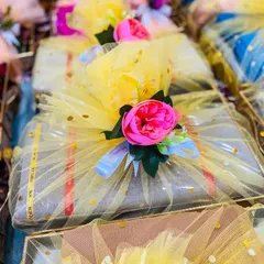 A collection of wedding favours wrapped in bright yellow tulle. Each package is adorned with a vibrant pink peony, making for a cheerful and eye-catching gift for guests at a mehandi or haldi ceremony.
