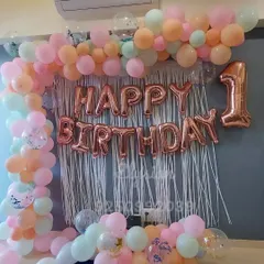 A beautiful pastel-themed decoration for a first birthday at home. The wall is decorated with a white fringe curtain, rose gold "Happy Birthday" letters, and a balloon arch in soft pastel shades.