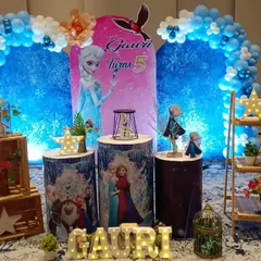 This Disney Frozen themed party for a fifth birthday features a circular balloon arch in shades of blue and white. The stage includes character cutouts of Elsa and Anna, themed display stands, and illuminated letters for the name.