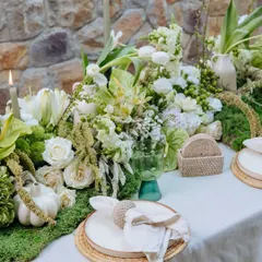 A close-up of our Earth Table setting. A crisp linen napkin is held by a woven ring on a ceramic plate, surrounded by a runner of moss and white flowers against our signature stone wall.
