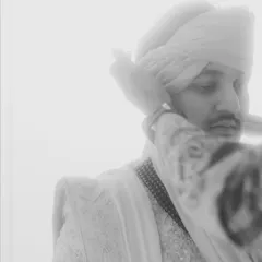 A soft, high-key black and white portrait of the groom adjusting his turban. The dreamy, ethereal quality of the light creates a moment of quiet contemplation.