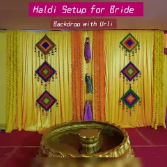 Here is a classic Haldi setup for the bride, featuring a large brass urli for the ceremony. The backdrop is made of rich yellow fabric, decorated with strings of marigolds and colorful, geometric hangings that add a festive touch.