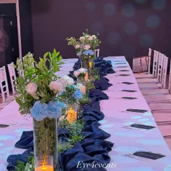 Here is a view of a long dining table decorated in a classic blue and white theme. Tall glass vases with blue and white floral arrangements run down the center, complemented by a navy blue fabric runner and the warm light of candles.