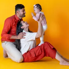 A fun and vibrant family photo against a bright yellow backdrop. The playful interaction between the parents and their baby girl makes for a joyful and energetic portrait.