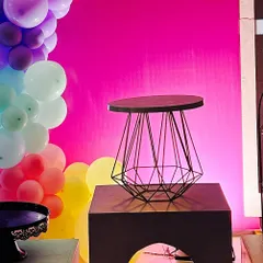 A modern geometric wire table stands on a pedestal, set against a bright pink wall and a rainbow balloon garland.