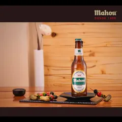 This image showcases a Mahou Clasica bottle with assorted skewers on a slate platter. The setup is designed to highlight the beer as a perfect companion for appetizers and social gatherings.