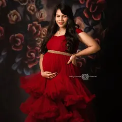 A vibrant portrait of a mom-to-be in a stunning, multi-layered red tulle gown. She stands against a dark, painted floral backdrop, creating a rich and romantic fine art maternity look.