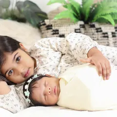 A beautiful portrait of sisterly love. The older sister lies protectively beside her sleeping newborn sibling, looking right at the camera, creating a sweet and engaging photo.