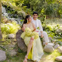 A classic couple portrait set in a lush, green garden with a waterfall. Their coordinated designer outfits in yellow and white add a touch of sunshine to this serene and romantic pre-wedding picture.