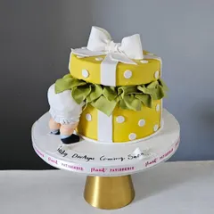 A playful and adorable baby shower cake designed to look like a gift box. The cake features a cute baby bottom peeking out, a big white bow, and a cheerful yellow polka dot pattern.