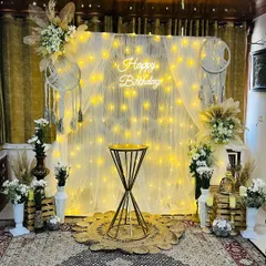 A beautiful boho-themed birthday corner set up in a living room, using a fairy light curtain, macrame dreamcatchers, and pampas grass.