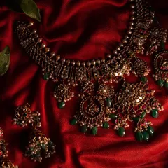 A close-up of an exquisite antique-finish temple jewelry set, with intricate details and green stones, resting on a rich red silk cloth.