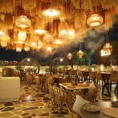 The interior of Sesh Delhi, designed to capture the vibrant, bohemian spirit of Goa. The ceiling is a dense tapestry of straw and woven pendant lights, creating a dramatic, immersive experience complemented by natural stone flooring and rattan furniture.