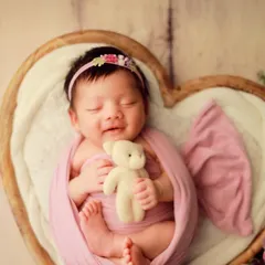 That fleeting, precious newborn smile. We live for these moments of pure joy. This little one is settled comfortably in our heart-shaped wooden prop, wrapped in a soft pink swaddle for a classic and heartwarming portrait.