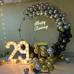 A modern and stylish 29th birthday setup. The decor features a circular balloon frame in black, silver, and gold, a 'Happy Birthday' neon sign, and large marquee numbers.