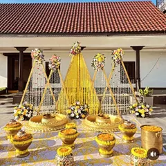 This is a detailed look at a Haldi ceremony setup with a backdrop of triangular wooden frames adorned with marigold and jasmine flower strings. The foreground has decorated pots (matkas) filled with flower petals.