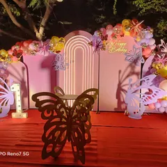 This is a beautiful butterfly-themed birthday setup for an outdoor evening party. It features pink arched backdrops, a custom butterfly-shaped table, and a colorful balloon garland.