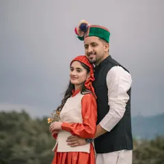 A classic portrait of the couple in full traditional Pahari dress, including the iconic Himachali cap. They stand together against a backdrop of green hills.