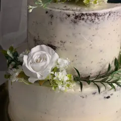 A detailed close-up of my rustic semi-naked cake. You can see the texture of the light buttercream coating over the dark cake, complemented by the delicate arrangement of food-safe white flowers and greenery.