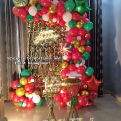 A festive birthday decoration featuring a gold shimmer wall, a 'Happy Birthday' neon sign, and a colorful red, green, and gold balloon arch.