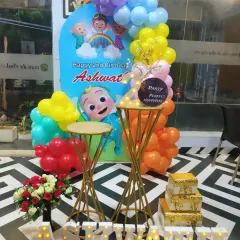 A bright and colorful Cocomelon theme for a 2nd birthday party. This setup features a custom character backdrop, a vibrant multi-colored balloon garland, and light-up letters spelling the birthday boy's name, 'ASHWATH'.