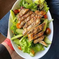 A healthy and satisfying grilled chicken salad. Sliced, herb-marinated chicken breast is served over a bed of fresh lettuce, cucumbers, and tomatoes, perfect for a light lunch or a healthy event menu.