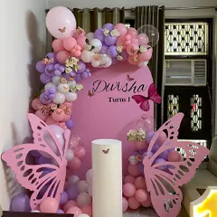 A beautiful butterfly-themed first birthday party in shades of pink and purple. The decor features a custom pink backdrop, an organic balloon arch, and large, elegant butterfly wing cutouts that create a magical photo corner.