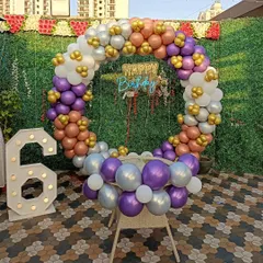 An outdoor birthday setup with a multi-color balloon ring against a green wall. The decoration includes a 'Happy Birthday' neon sign and a large light-up number '6'.