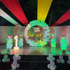 A large stage setup for an outdoor first birthday party. The decor includes a colorful fabric canopy, a balloon ring backdrop, and light-up balloon columns.