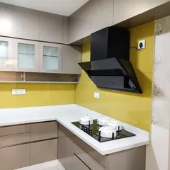 A contemporary L-shaped kitchen featuring neutral beige cabinets and a bright yellow lacquered glass backsplash that adds a pop of color. The design includes a black chimney and frosted glass upper cabinets for a clean and modern look.