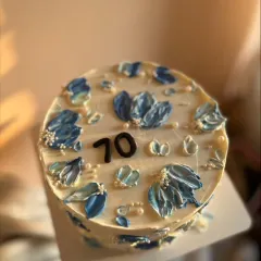 A close-up of a 70th birthday cake decorated with blue and white palette knife flowers. The chocolate sponge is filled with salted caramel and covered in vanilla buttercream.