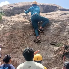 A participant in full gear practices his ascent on a large boulder in Ramanagara, with fellow campers watching and learning.