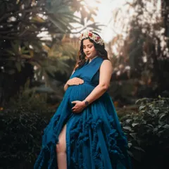 A close-up portrait of a mother-to-be in a lush green setting, wearing a deep blue gown and a floral crown. Her serene expression captures a moment of quiet reflection.