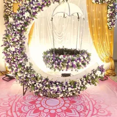 I create elegant stages for naming ceremonies, like this one with a crescent moon floral frame. The cradle is also decorated with matching purple and white flowers, and the backdrop uses sheer and gold curtains for a traditional and beautiful seemantham event.
