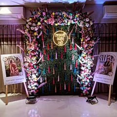 A festive backdrop for a Navanu Tapasvi event. The design includes a dense floral arch over a green wall with colorful tassels and personalized photo stands on either side.
