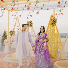 A couple walks hand-in-hand through their beautifully decorated haldi venue in the Dehradun hills. The vibrant purple and yellow decor creates a stunning visual against the mountain backdrop.