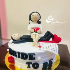 A playful 'Bride to Be' cake for a bachelorette party or bridal shower. The handcrafted fondant figures show the bride-to-be relaxing while her groom lies down, with a calendar marking the big day.
