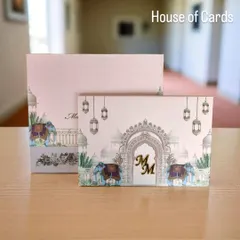 This pastel pink invitation features a palace-inspired design with elephants and hanging lanterns, complete with a custom gold monogram for a personal touch.