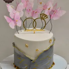 A beautiful two-tier engagement cake in a grey and white marble design with gold leaf accents. The top is decorated with delicate pink wafer paper butterflies and a golden ring topper, making it both modern and romantic.