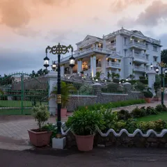 The entrance to 'The Palace Promised' at dusk. The warm lighting accentuates the classical columns and ornate details, while the traditional lamp posts illuminate the winding driveway and lush gardens.
